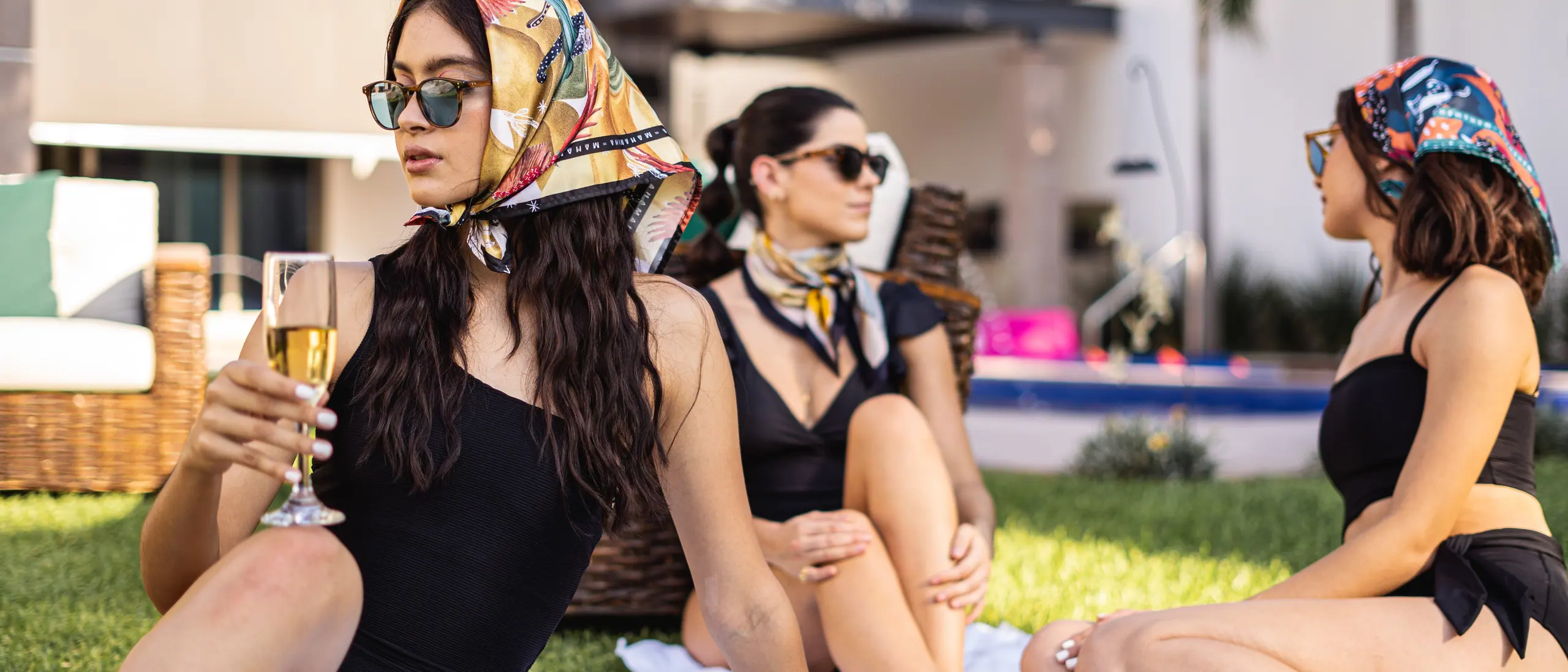Tres mujeres con pañuelos de seda y lentes de sol, brindando sobre el pasto junto a la alberca